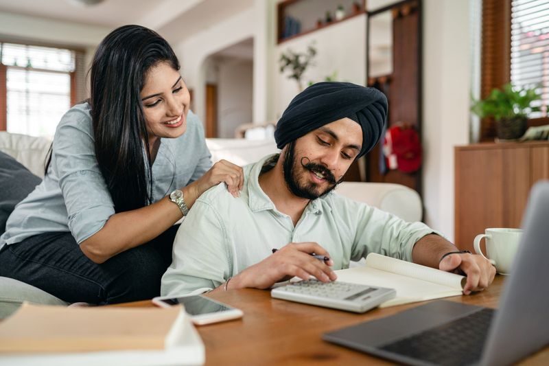 Smiling young Indian female in casual clothes supporting husband during remote work at home sitting in front of laptop with calculator notebook smartphone and cup of coffee at table