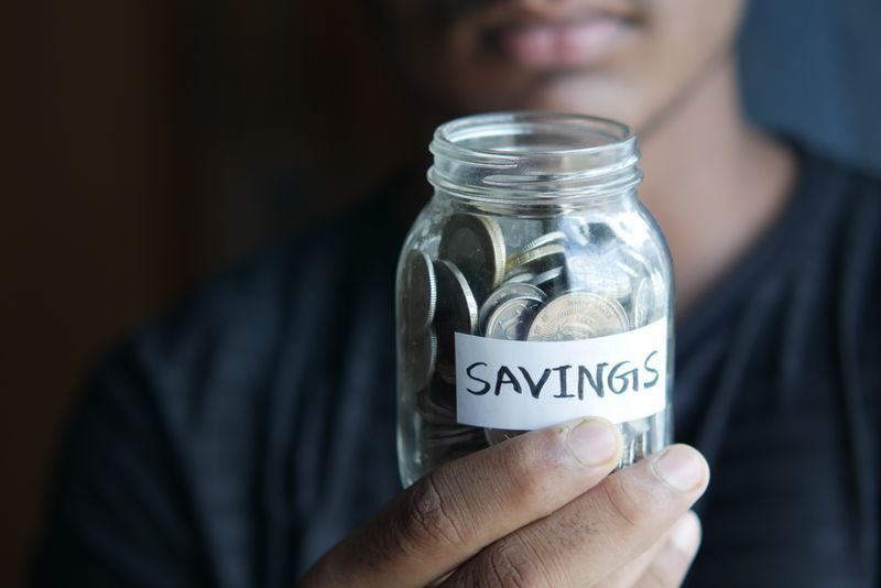 A close-up image of a person's hand holding a jar full of coins labeled 'Savings'.