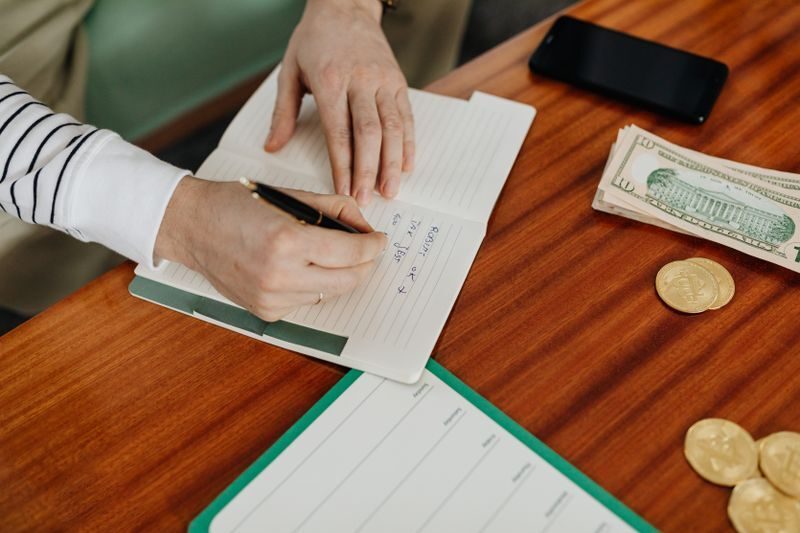 Person writing in a notebook on a wooden table with cash, coins, and a phone nearby.