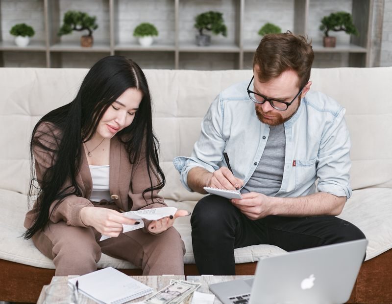 A man and woman sitting indoors, reviewing expenses with notebooks and money on the table.